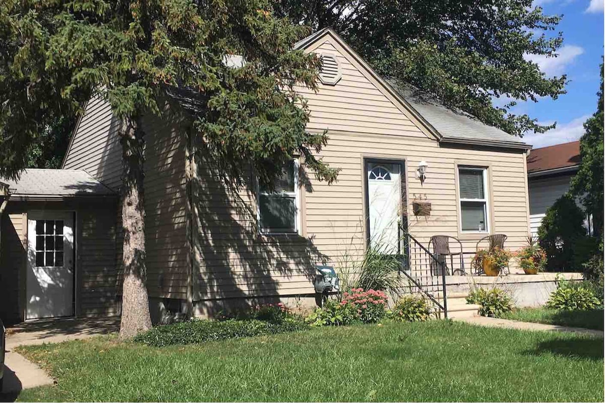 A modern bungalow is depicted with light brown siding and a welcoming entrance. A set of steps leads to the front door, complemented by two chairs and a small table on the porch. Green grass and colorful flower beds enhance the outdoor appeal.