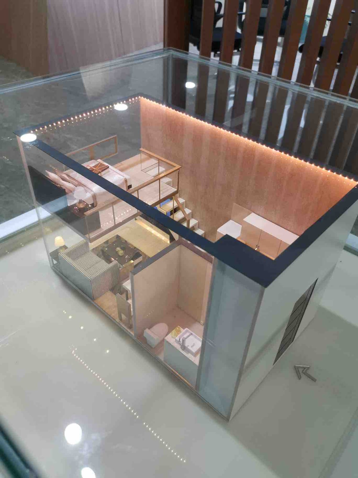 A scaled model of the loft-type condominium is displayed in a protective case. The interior layout is visible, showcasing a living area, kitchenette, staircase, and separate sleeping spaces. Soft lighting outlines the walls, enhancing the model's design features.