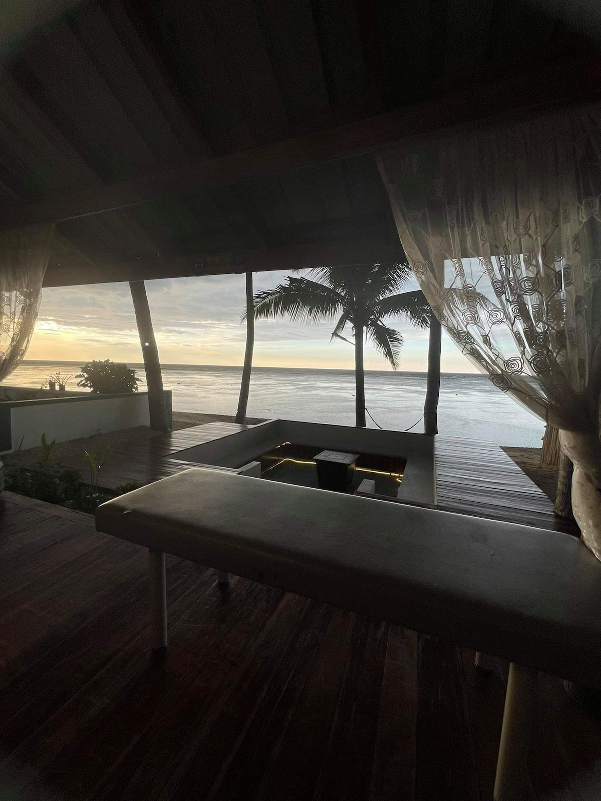 A tranquil beachfront view showcases soft sunset hues through large openings that frame the sea and palm trees. The wooden deck invites relaxation, while a massage table is positioned nearby, allowing guests to unwind while enjoying the serene environment.