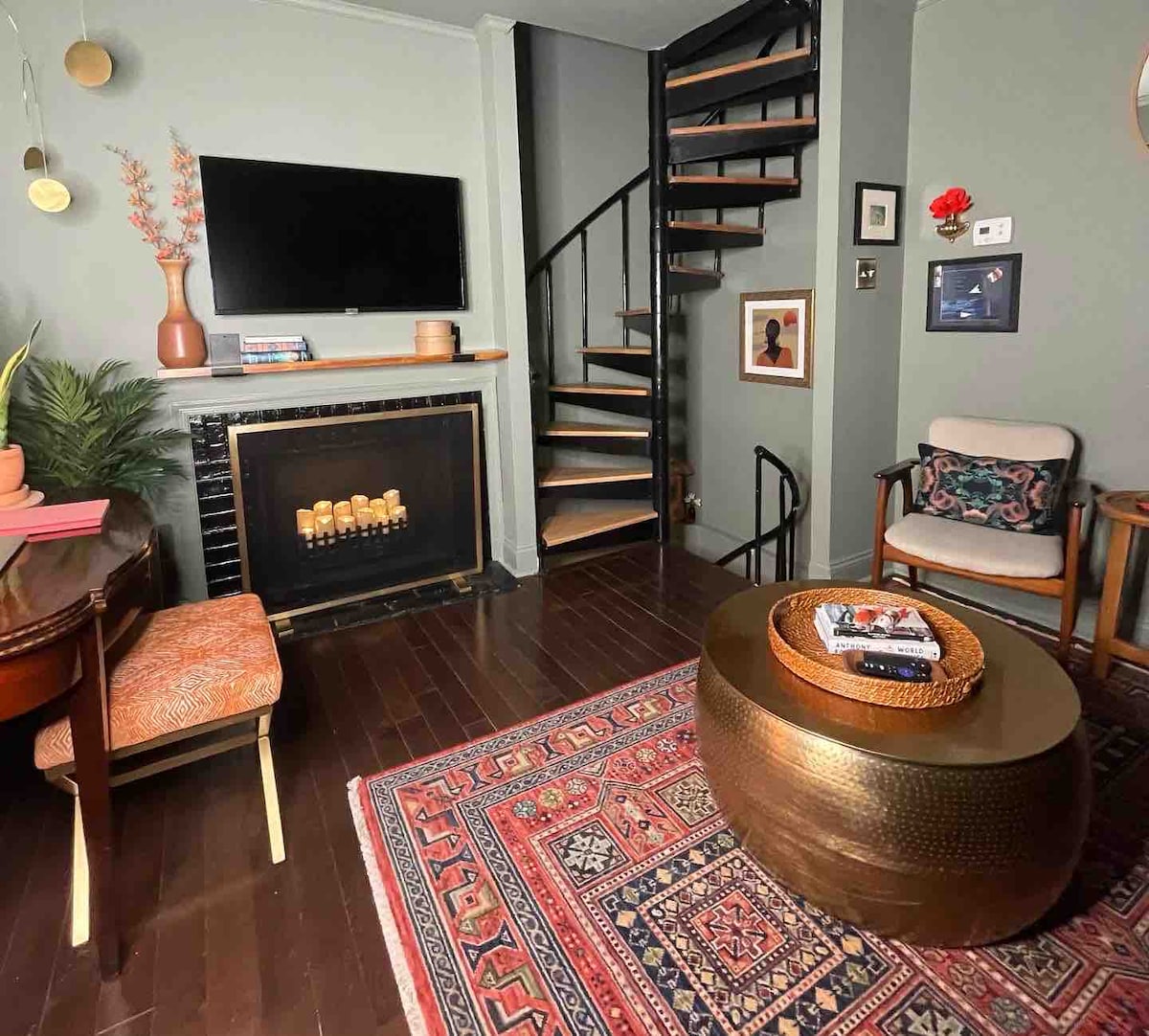 The living room features a stylish sofa and a decorative fireplace with an electric candelabra. A wall-mounted smart TV is included, and a spiral staircase leads to upper floors. The space is accented by a vibrant area rug and tasteful decor elements, creating a cozy environment.