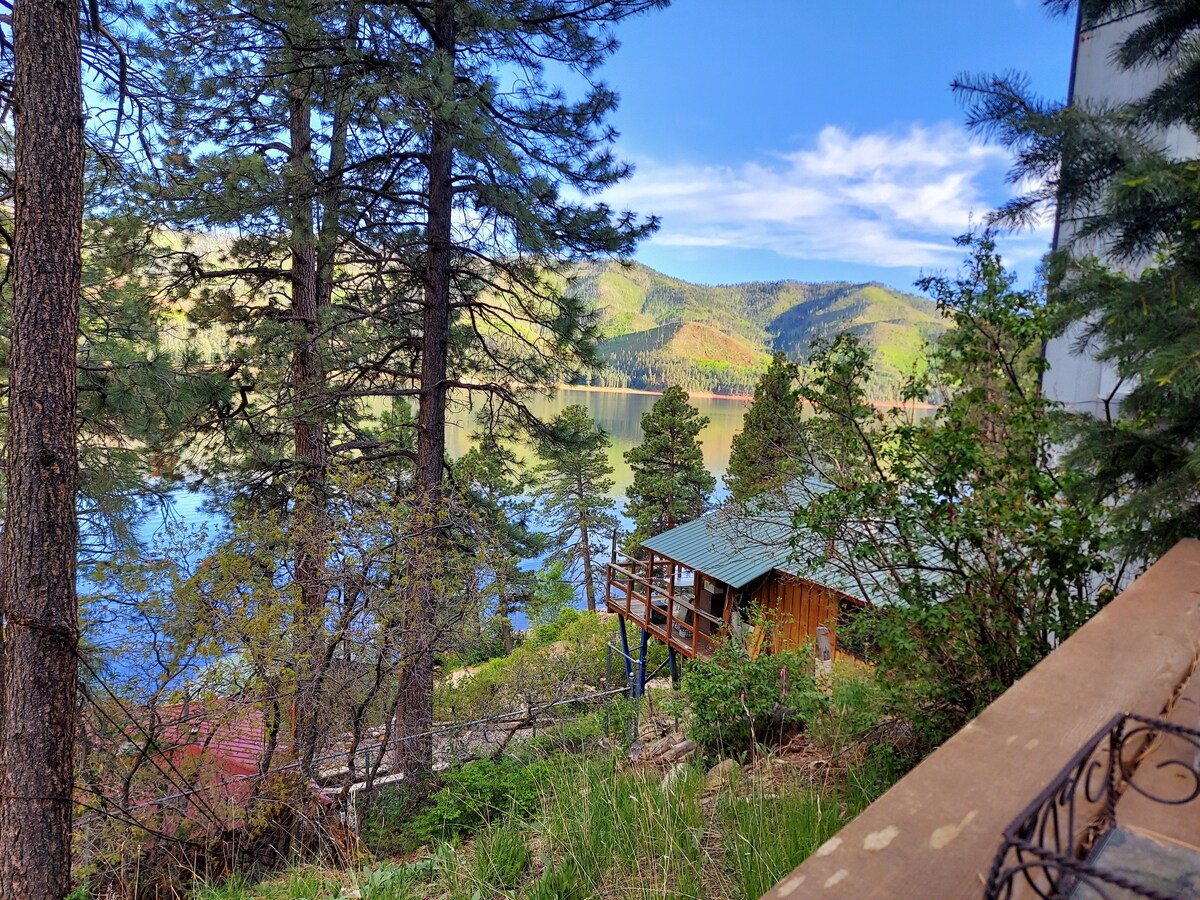 A tranquil lakeside view features a wooden cabin nestled among tall trees. The calm water reflects the surrounding mountains, while scattered greenery adds a natural touch to the landscape. Blue skies and soft clouds create a serene atmosphere, perfect for outdoor relaxation.