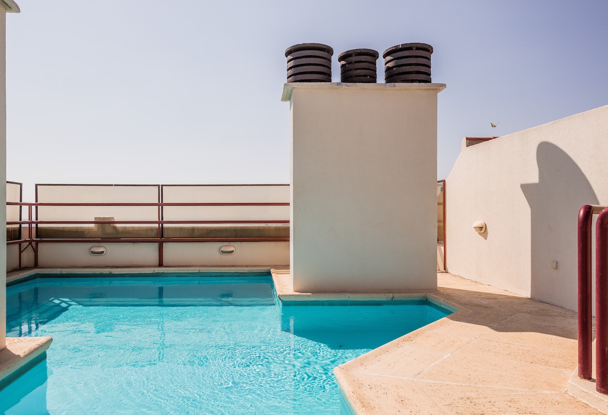 The rooftop pool area features a clear blue water pool surrounded by beige tiles. Large utility pipes are situated beside the pool, and a low wall provides a barrier with an unobstructed view of the sky above.