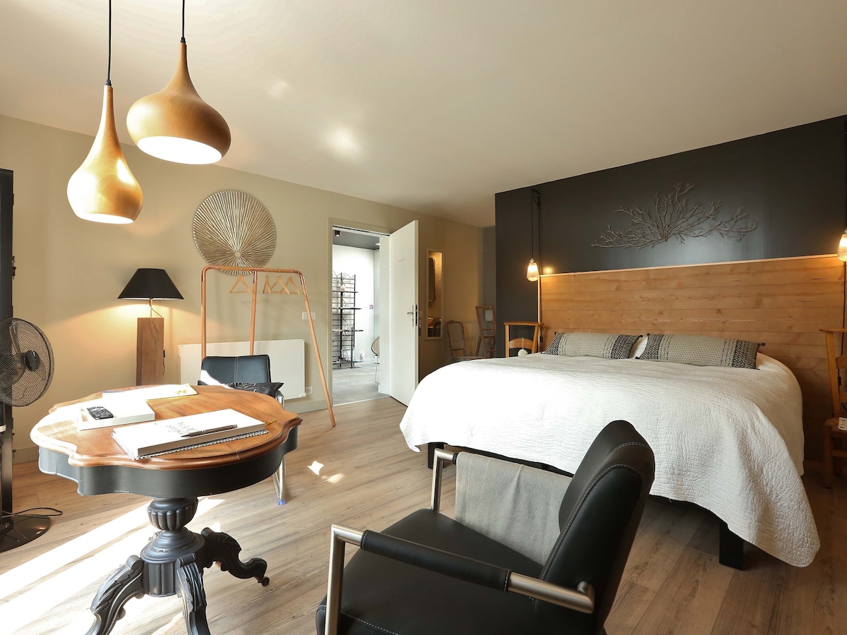 A spacious guest room features a large bed with a wooden headboard against a dark wall. Two pendant lights hang above, illuminating a stylish table and chair set. A fan and a clothing rack are visible, contributing to a calm and comfortable environment.
