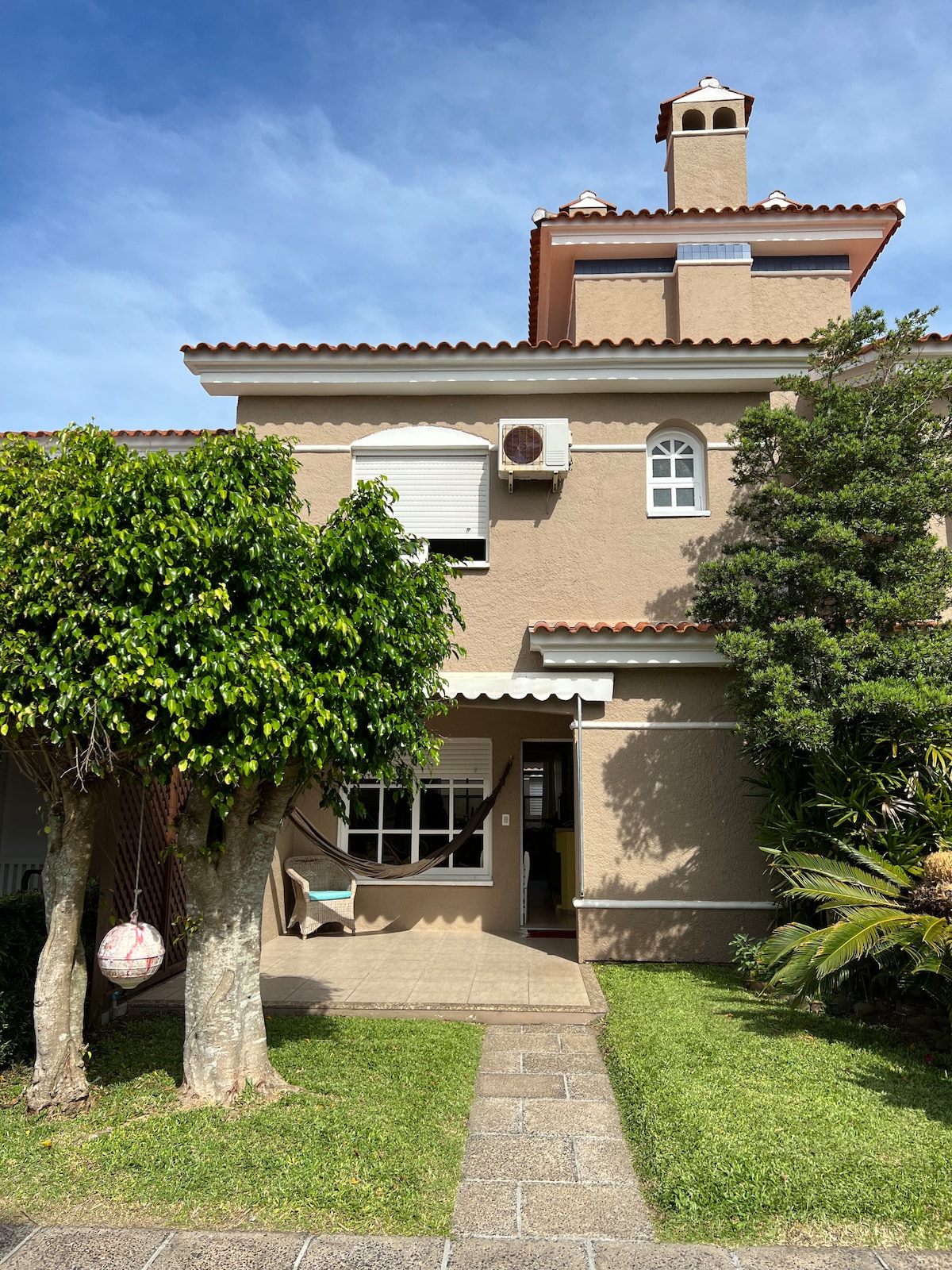 The exterior of the house is captured, featuring a two-story structure with a light brown façade and a tiled roof. Lush greenery surrounds the entrance, complemented by a swing hanging from a tree. Pathway tiles provide access to the front door, under a bright blue sky.