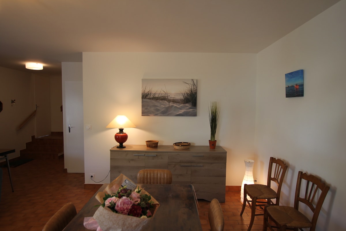 A dining area is showcased, featuring a wooden table set with a floral centerpiece and surrounded by chairs. A sideboard displays decorative items, with a lamp providing soft lighting. Two artworks adorn the walls, depicting serene beach scenes and adding to the room's tranquil ambiance.