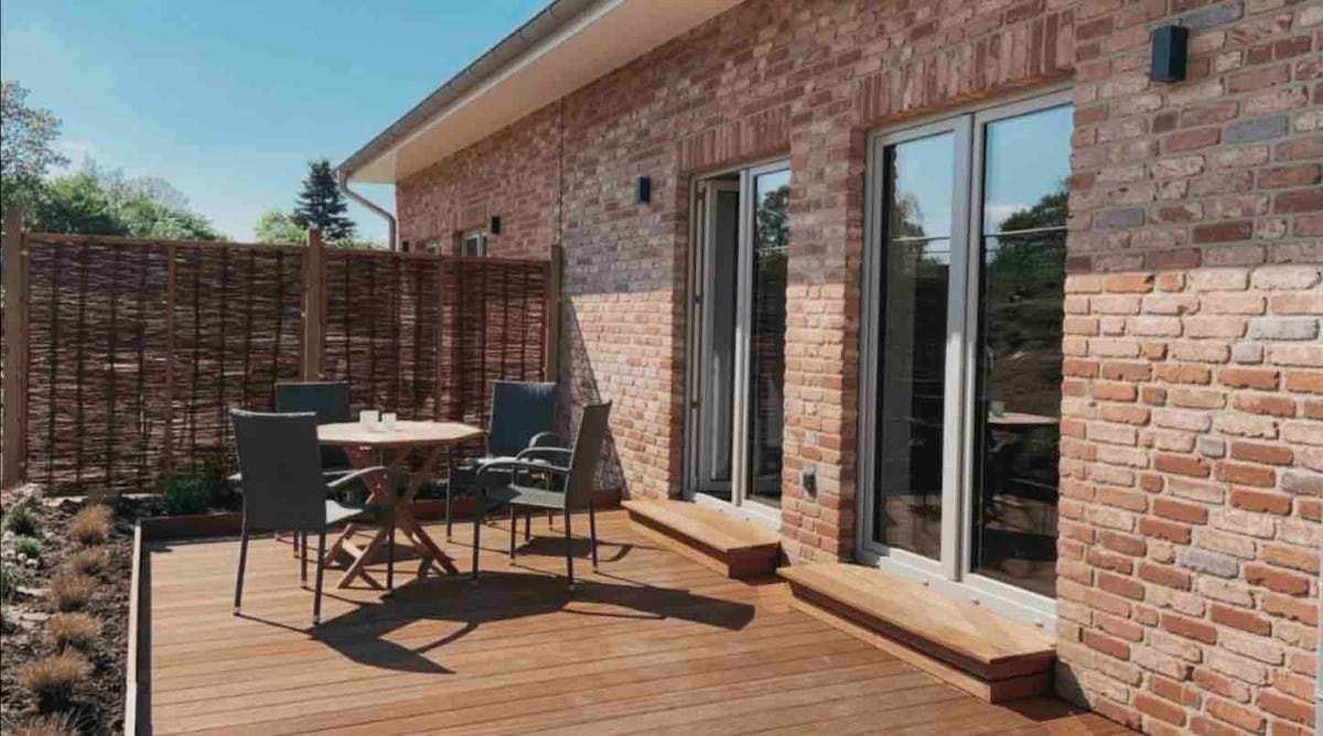 An outdoor terrace features wooden decking, complemented by a round dining table surrounded by four chairs. Large glass doors provide access to the interior, and a woven privacy screen offers separation from surrounding areas, creating a serene outdoor space.