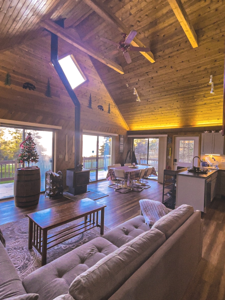 The interior features a vaulted wooden ceiling with exposed beams and expansive windows that invite natural light. A cozy seating area with a sofa is positioned near a rustic coffee table. A dining space and modern kitchen with stainless appliances are visible in the background.