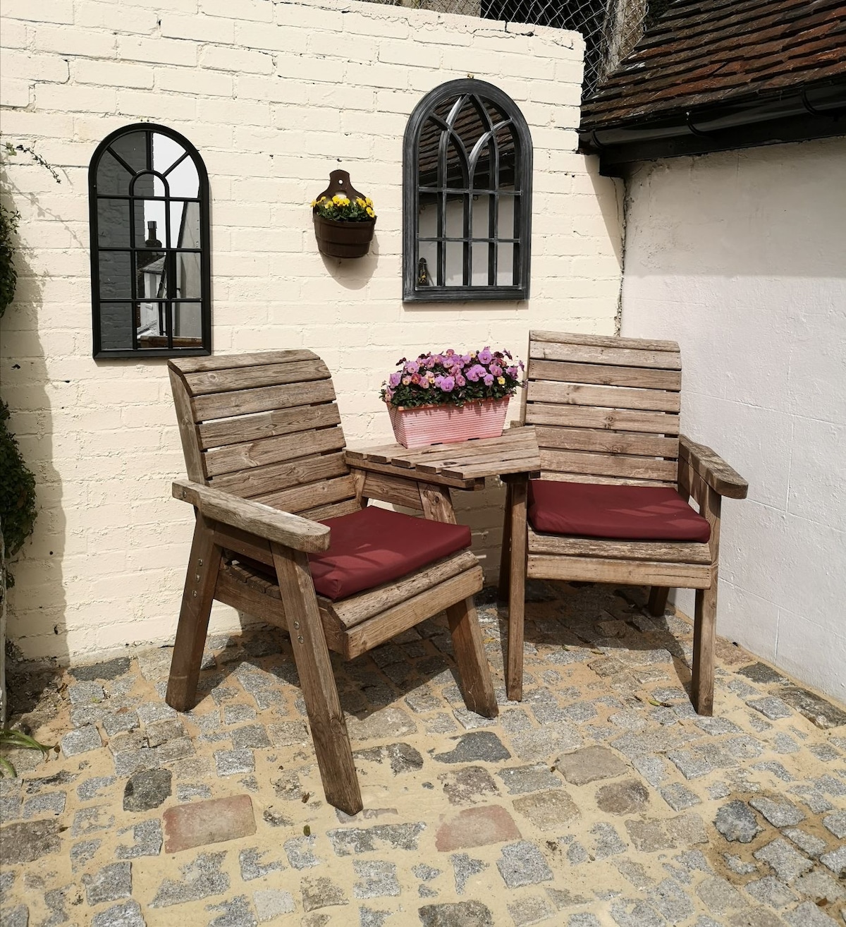 Two rustic wooden chairs with red cushions are positioned adjacent to a small wooden table, set against a cozy cottage wall. Above the chairs, a decorative wall planter filled with colorful flowers adds a touch of charm, while two arched windows reflect natural light.
