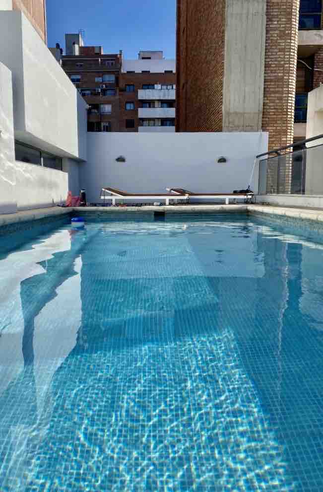 A swimming pool is featured on a rooftop, surrounded by clean white walls and a smooth deck. Sun loungers are positioned alongside the pool for relaxation. The clear blue water reflects sunlight, creating a refreshing ambiance in the city setting.