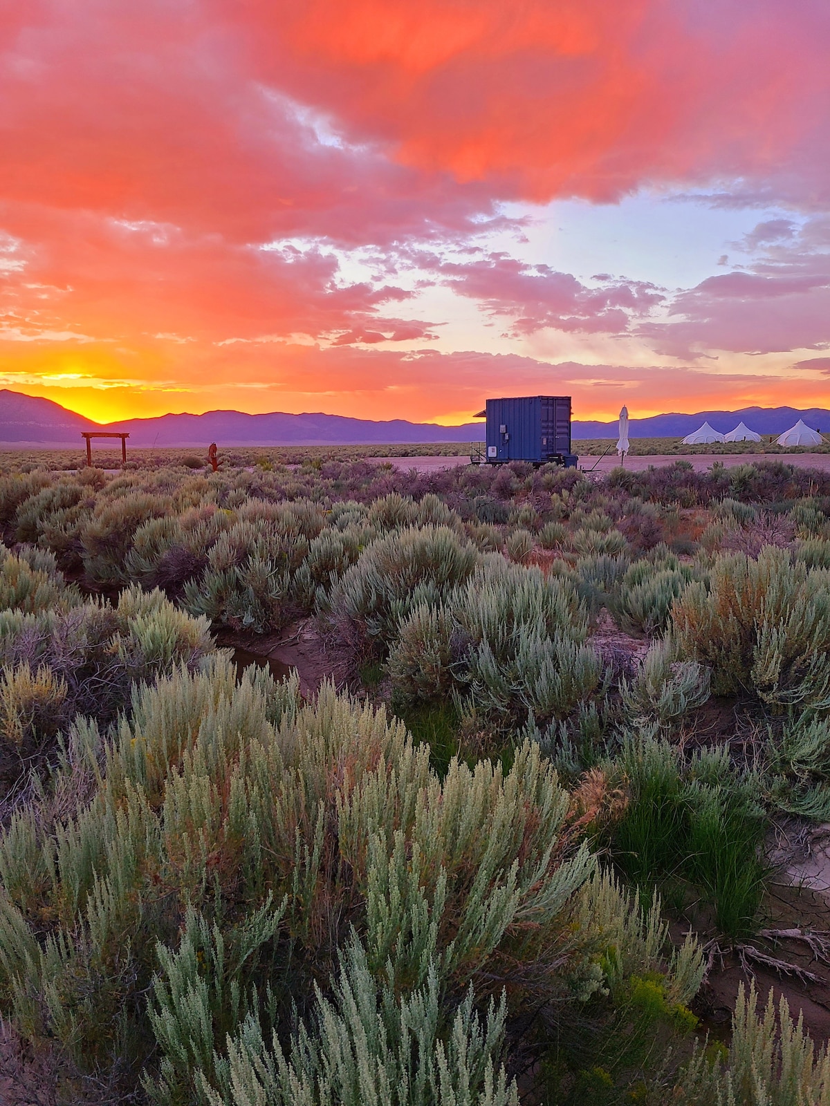 A serene landscape is captured during sunset, with colorful hues filling the sky. In the foreground, lush sagebrush surrounds a small structure, which is complemented by nearby tents. Mountain silhouettes are visible in the distance, enhancing the tranquil setting.