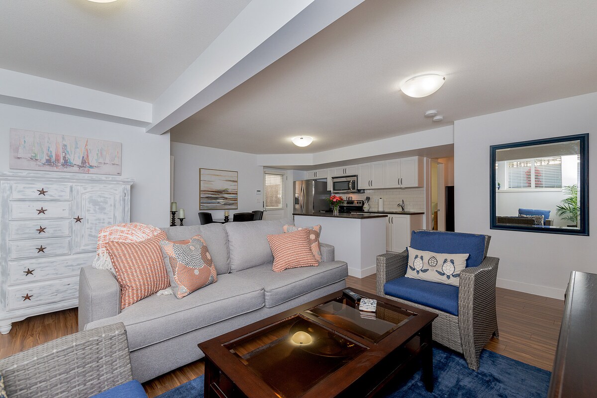 A spacious living area features a light gray sofa adorned with decorative pillows, complemented by a blue accent chair. A wooden coffee table sits in front, while a sleek kitchen with white cabinetry is visible in the background. Natural light is welcomed through a nearby window.