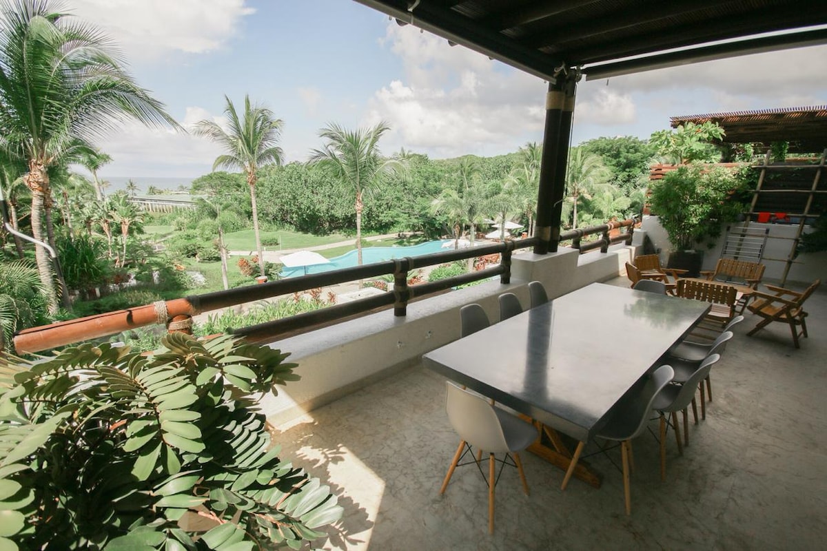 An outdoor dining area features a long gray table surrounded by white and wooden chairs. Lush greenery and palm trees are visible in the background, alongside a glimpse of a pool. The space is covered for shade, providing comfort for outdoor meals.