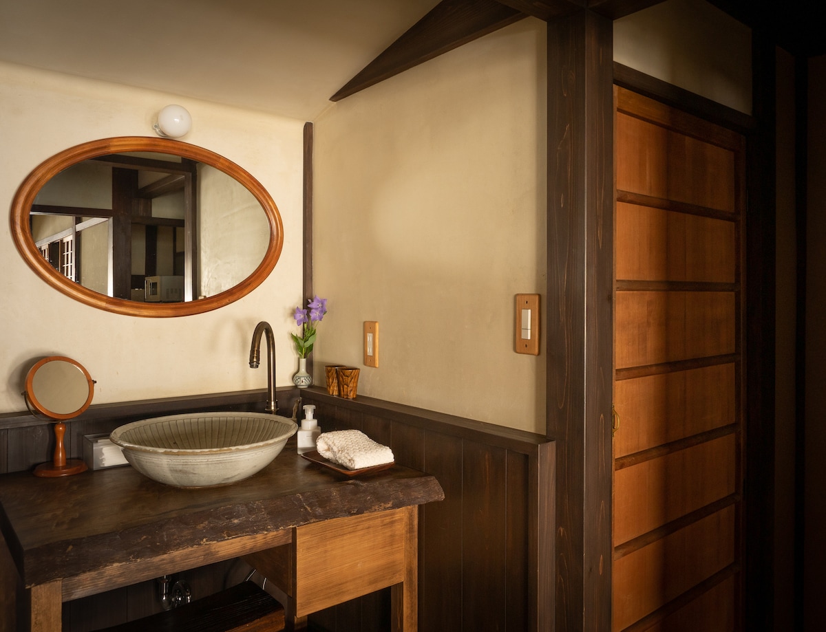 A wooden washstand is displayed with a round basin and a decorative mirror above. A small vase with flowers adds a touch of nature. A wooden door is partially visible, showcasing the natural materials used in the design.