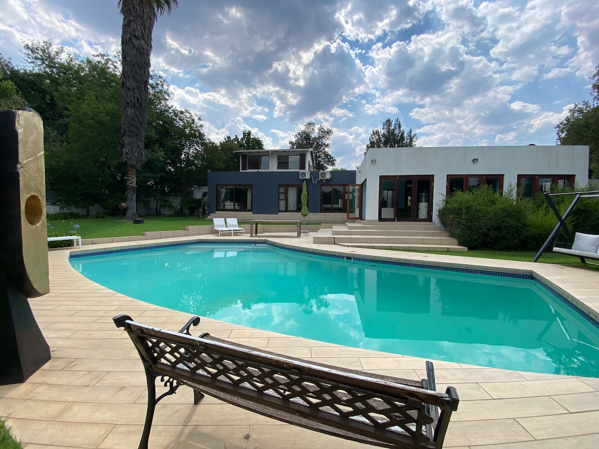 A spacious outdoor area features a large pool surrounded by a tiled deck. Lounge chairs are positioned nearby, and a sculptural feature adds visual interest. The house is visible in the background, framed by greenery and a partly cloudy sky.