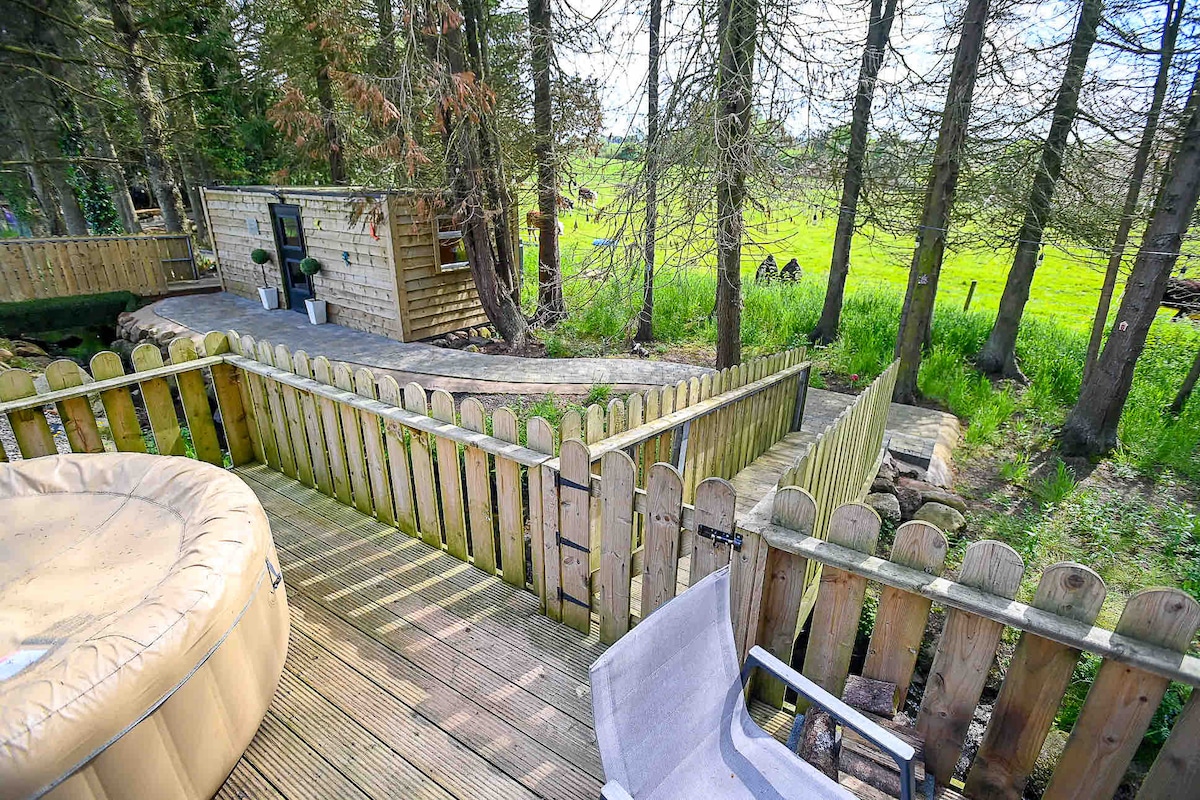 A wooden deck surrounds a private hot tub, framed by a wooden fence. A pathway leads to a cozy cabin set among trees, with a lush green landscape visible in the background. The scene captures a tranquil outdoor setting.
