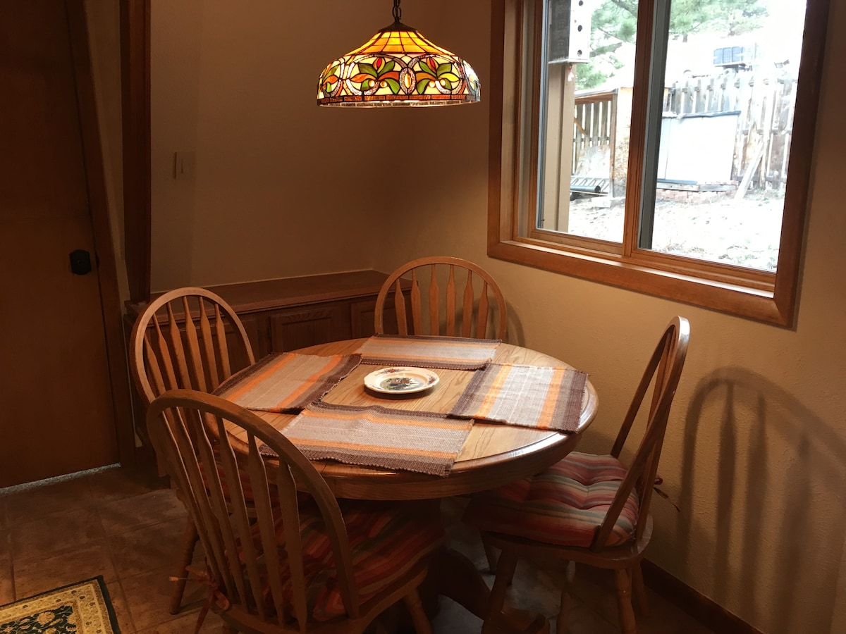 A round wooden dining table with four matching chairs is highlighted, featuring colorful cushions. Above, a stained glass pendant light adds character. Natural light enters through a nearby window, which overlooks the outdoor space.