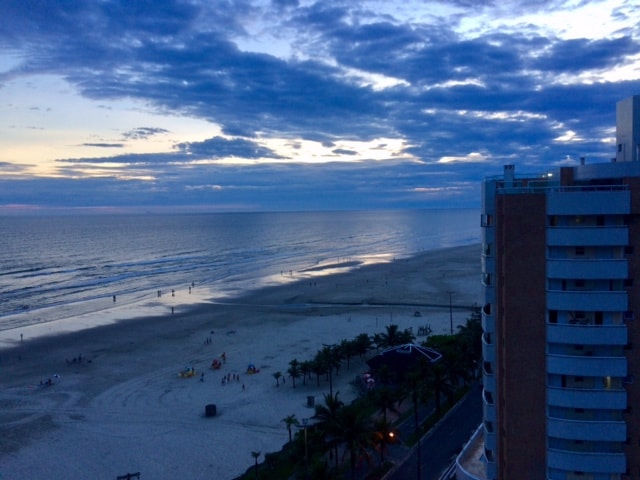 A serene beach view captures the gentle waves meeting the shore under a soft twilight sky. The sandy beach is lined with small figures, and faint outlines of palm trees are visible along the coastline, creating a calm atmosphere.
