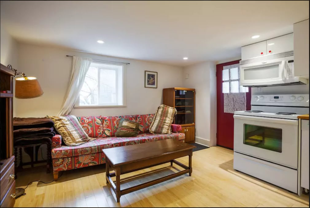 A well-lit living area features a colorful patterned sofa paired with a wooden coffee table. A window allows natural light to brighten the space, while a red door adds a pop of color. Shelving units are positioned nearby for additional storage.
