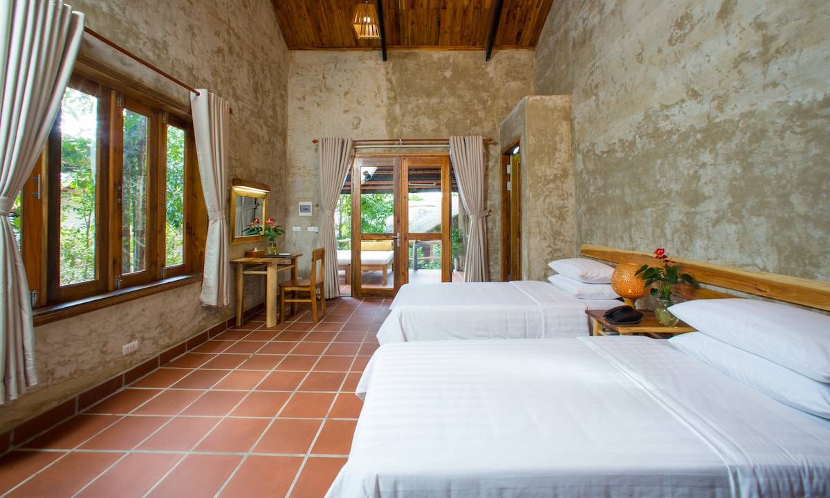 A tranquil room features two spacious beds with simple white linens. Natural light enters through large windows adorned with light curtains. A wooden desk and chair complement the rustic decor, while plants add a touch of greenery to the space. A door leads to an outdoor view.