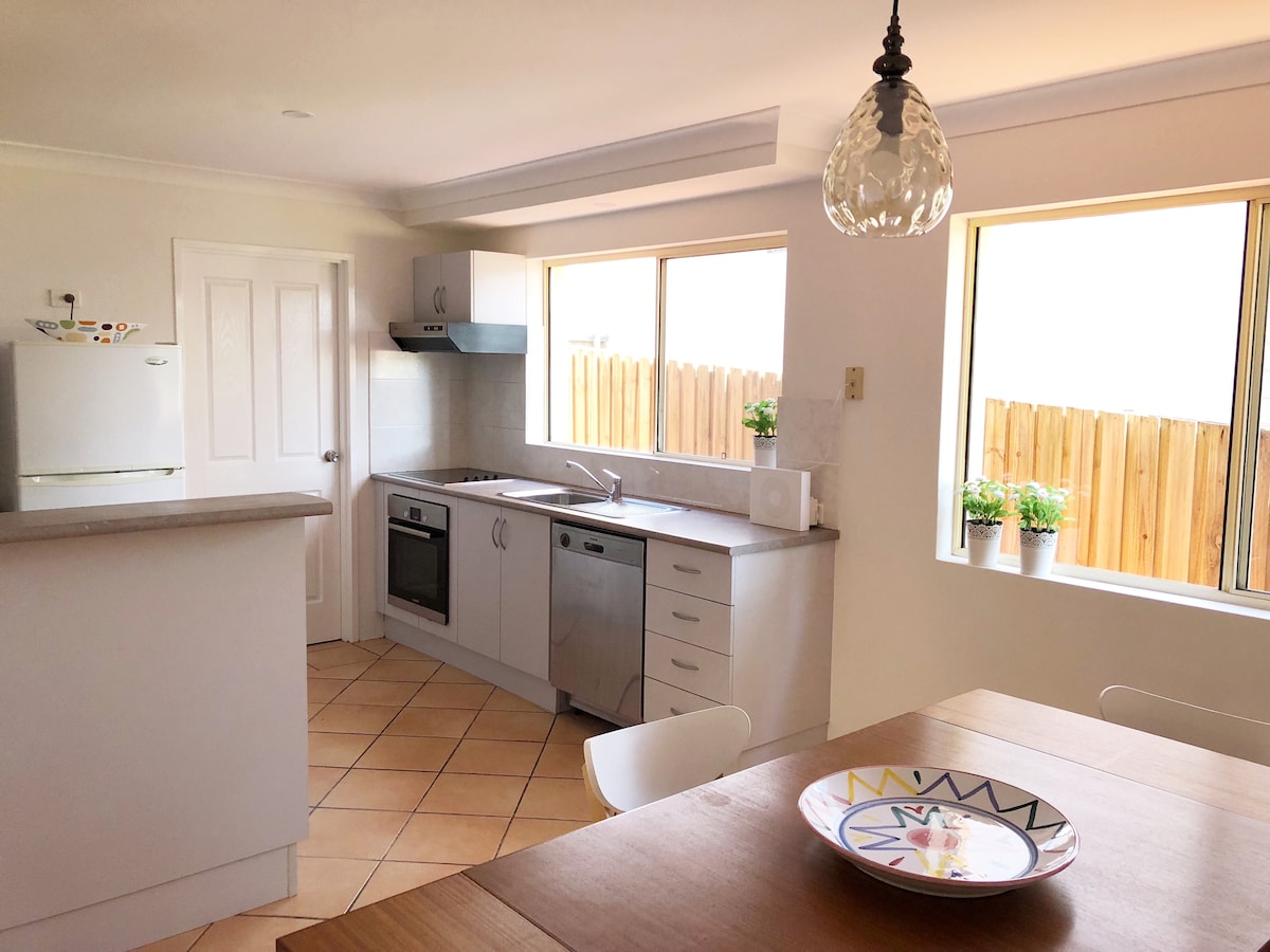 A well-lit kitchen area features modern appliances, including an oven and dishwasher, set against light cabinetry. Natural light floods in through two large windows, with green potted plants placed on the sill. A wooden dining table is positioned nearby, complemented by a decorative plate.