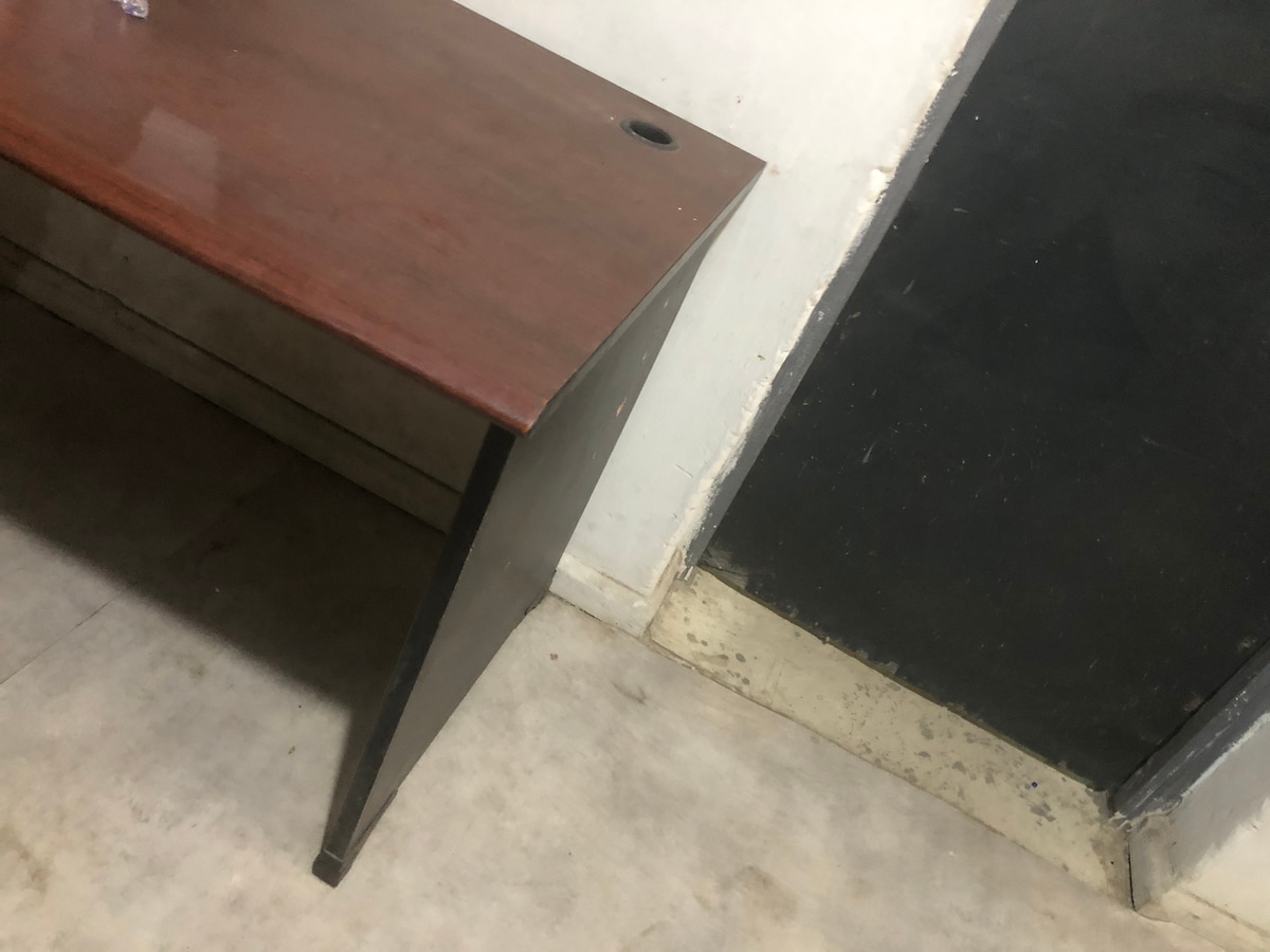 A simple wooden desk is positioned against a wall, featuring a smooth surface and an opening for cable management. The floor is lined with light-colored tiles, and the image captures a corner where the desk meets the wall.