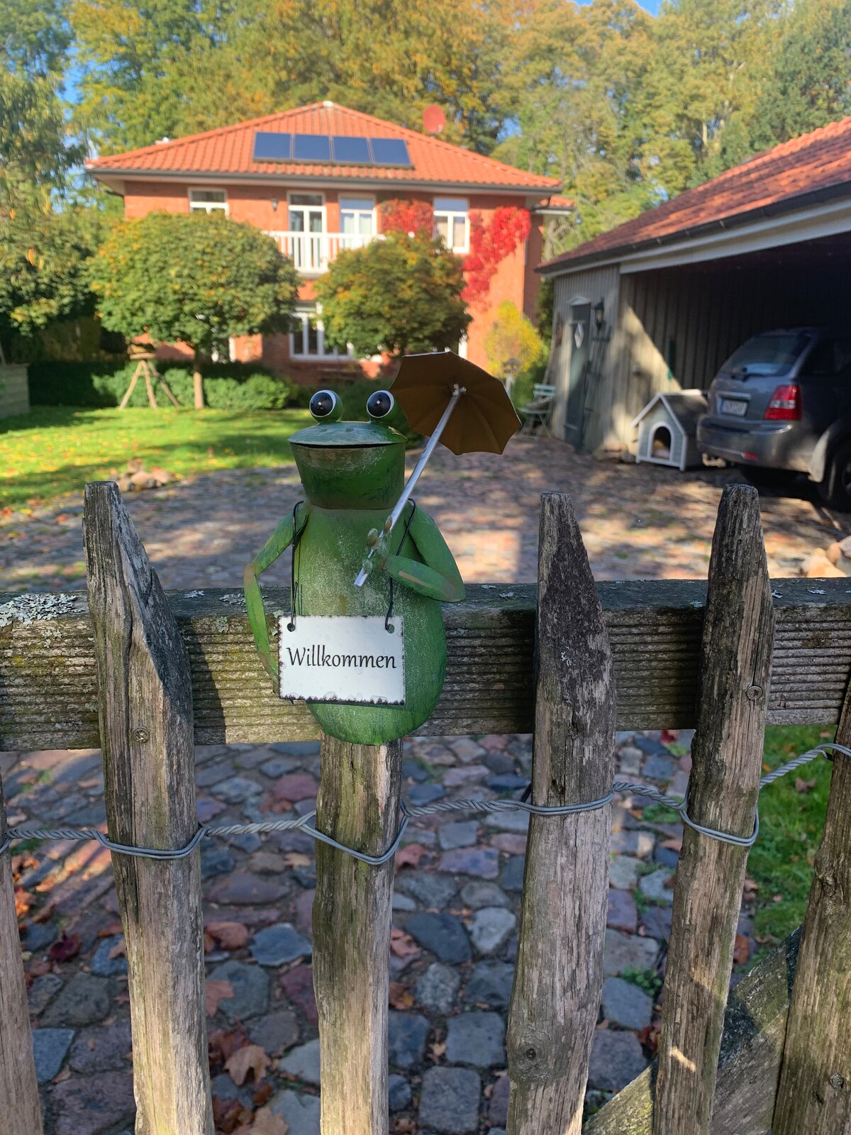 A whimsical frog figure holding an umbrella stands on a wooden fence, welcoming visitors with a small sign. The background features a charming house surrounded by greenery and a peaceful driveway, enhancing the inviting atmosphere of the property.