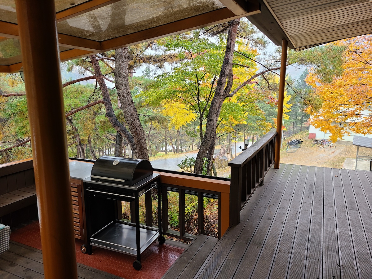An outdoor terrace overlooks a scenic area, framed by trees displaying autumn colors. A barbecue grill is positioned on the wooden deck, ready for use. The surrounding space features a well-maintained wooden surface, creating a relaxing environment for outdoor dining.