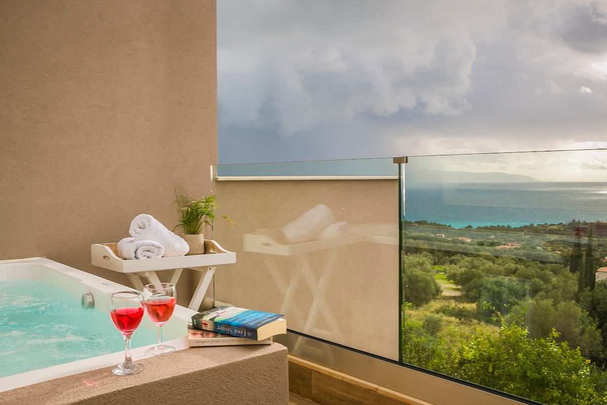A private Jacuzzi is positioned on the balcony, accompanied by two glasses of red beverage and a stack of books. Soft, rolled towels rest on a nearby tray. The panoramic view reveals lush greenery and the deep blue sea under gentle cloud cover.
