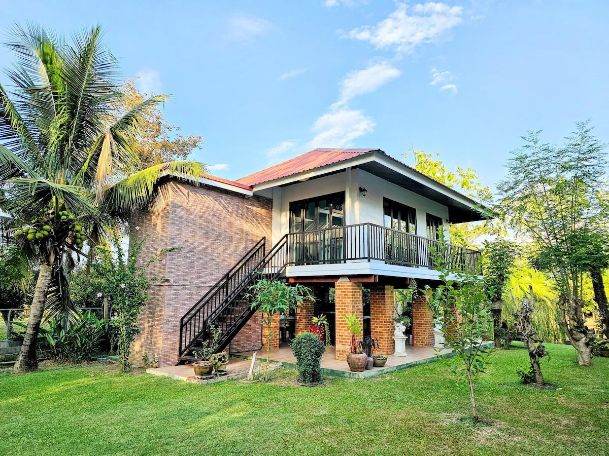 A two-story guesthouse is surrounded by greenery, featuring a combination of brick and wooden design. A staircase leads to the upper level, where large windows allow natural light to fill the space. Tropical plants and well-maintained gardens enhance the serene outdoor environment.