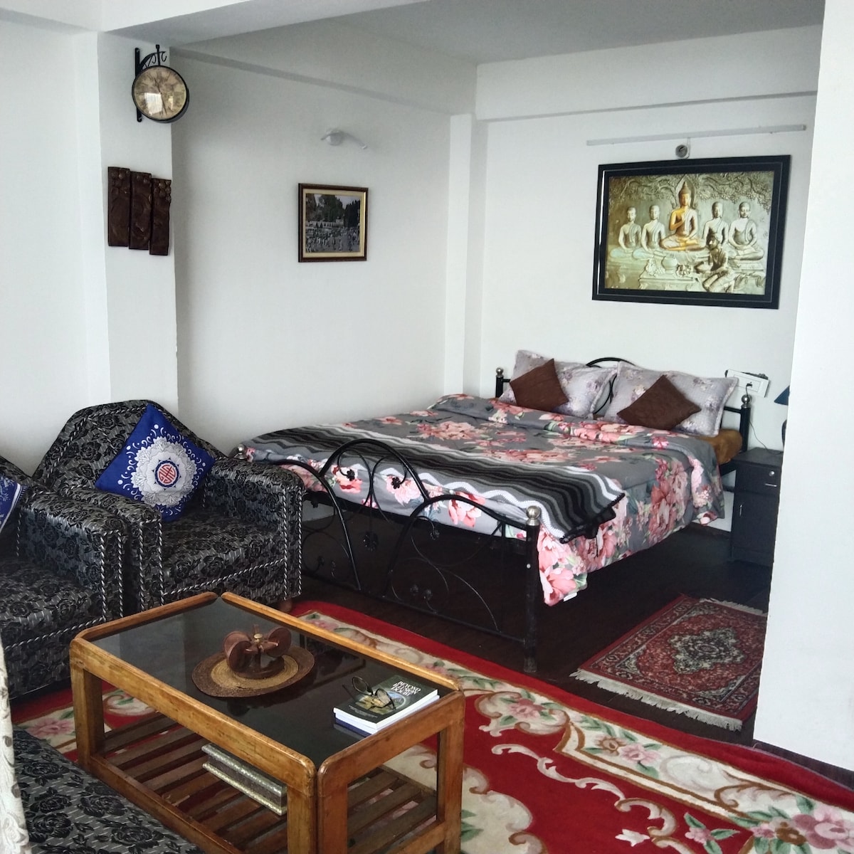A private suite features a spacious bed adorned with floral bedding, complemented by decorative pillows. A cozy seating area includes two patterned chairs and a wooden coffee table. Artwork depicting serene themes hangs on the wall, while a clock adds a timeless touch to the space.