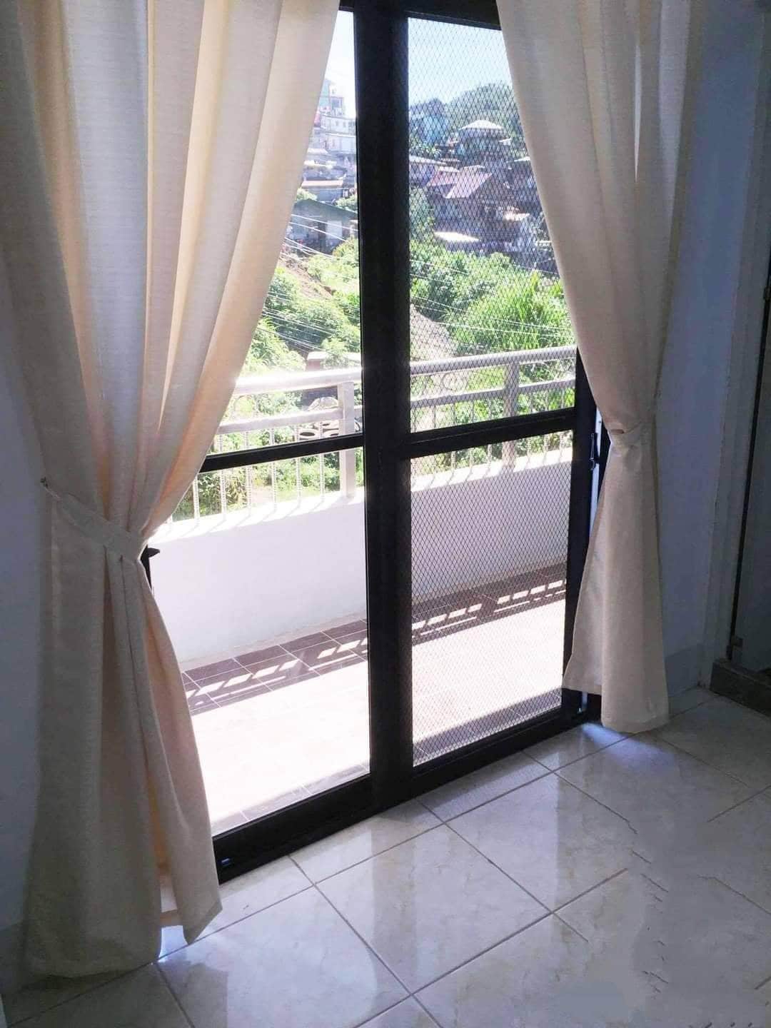 A sliding glass door, framed by beige curtains, leads to a balcony. Scenic views of rolling hills and buildings in the distance are visible. Light marble floors extend under the door, enhancing the airy space. Sunlight filters through the curtains, creating a bright atmosphere.