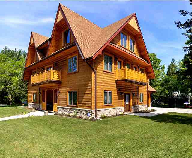 The exterior of the two-story wooden chalet showcases a blend of natural wood and stone finishes. Multiple balconies and large windows enhance the design. Lush green grass surrounds the home, providing a spacious outdoor area, while a basketball hoop is visible in the background.