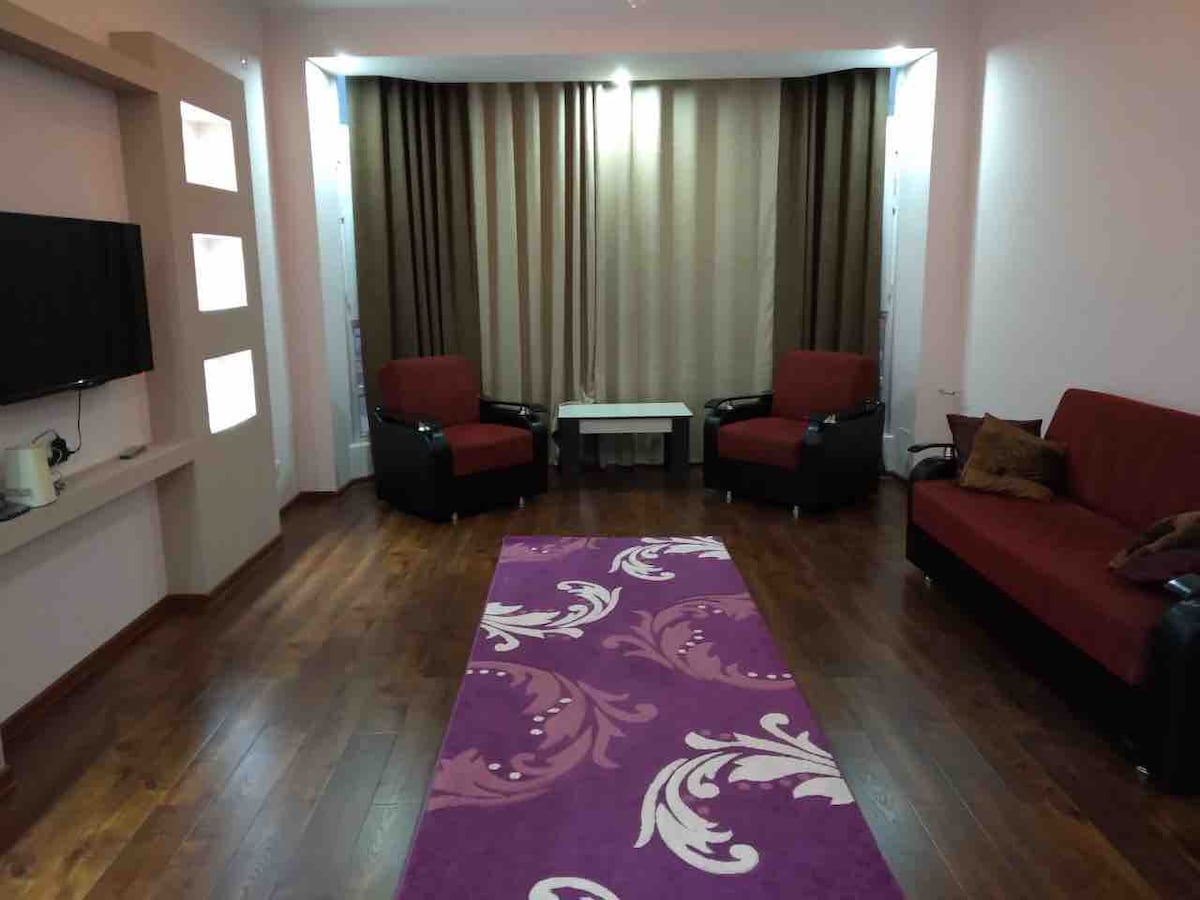 A spacious living area features two red armchairs and a black sofa, arranged around a small white coffee table. A vibrant purple rug with intricate patterns lies on the polished wooden floor. Floor-to-ceiling curtains in brown complement the natural light filtering in through the windows.