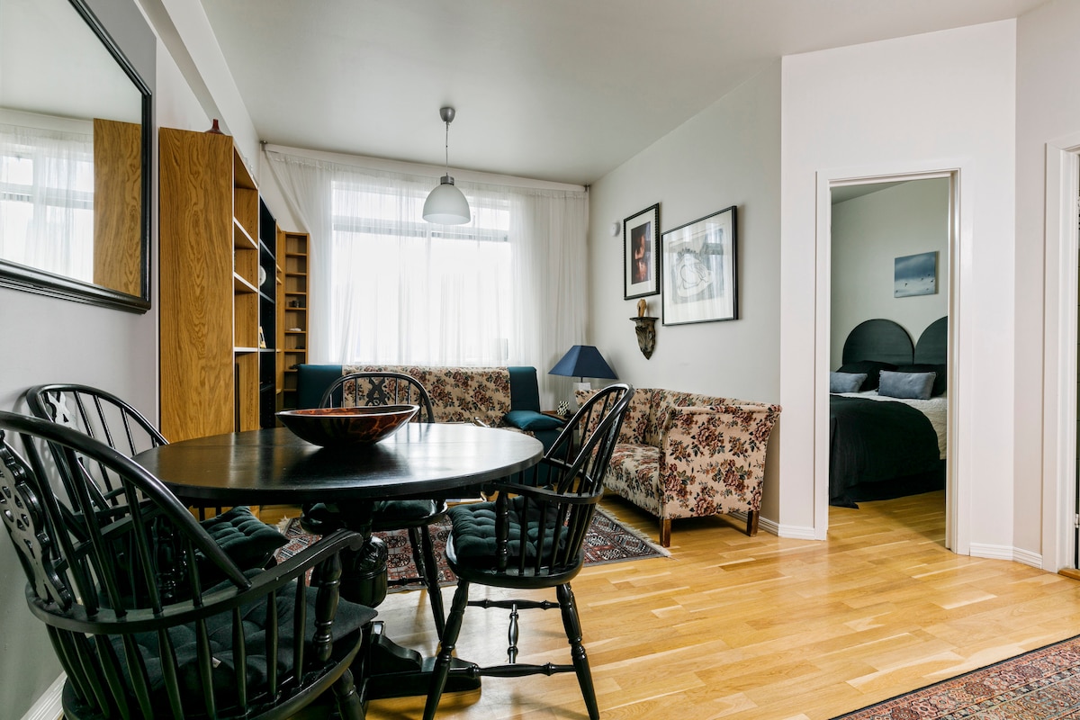 A dining area features a round table surrounded by four black wooden chairs. A floral-patterned sofa is positioned alongside a blue couch. Light enters through large windows, and a doorway leads to the bedroom, which is visible in the background.