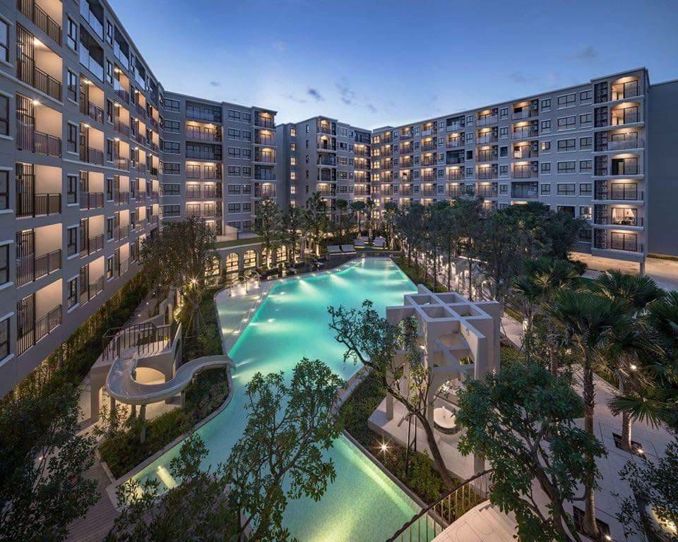 A spacious outdoor area features a large swimming pool surrounded by lush greenery and palm trees. The pool's turquoise water is contrasted by elegant lounge areas and a slide, with buildings framing the scene under an evening sky.