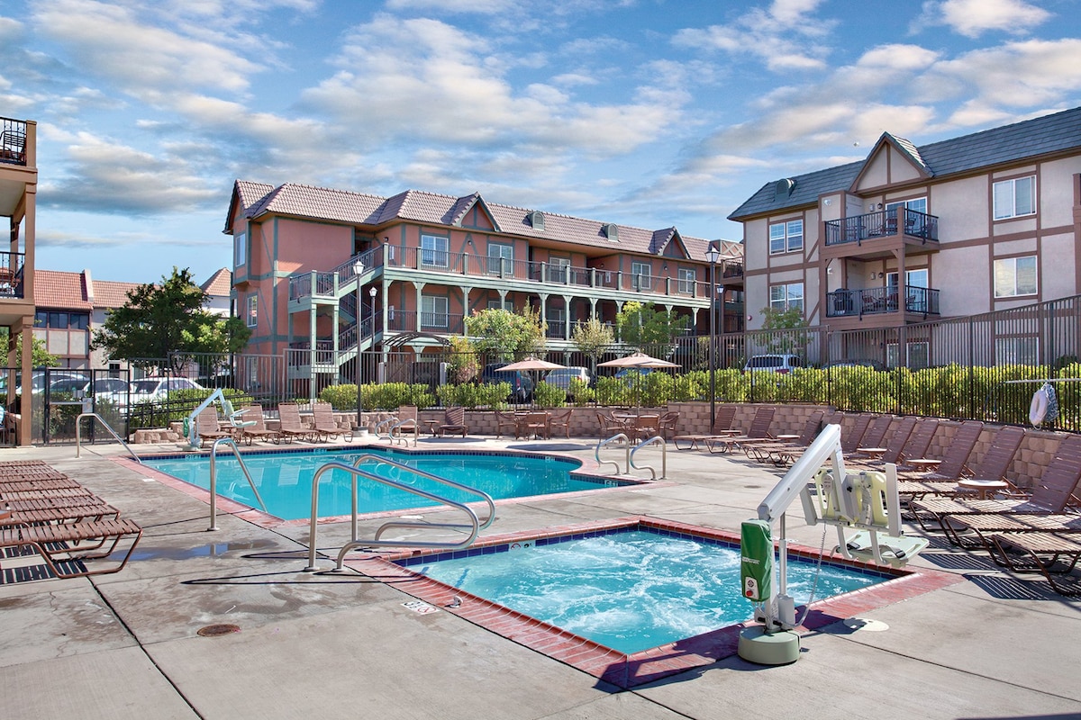 A spacious outdoor area features a swimming pool and a hot tub, surrounded by lounge chairs. Two brightly colored buildings are visible in the background, with shaded umbrellas providing additional comfort. The scene is complemented by green landscaping and a clear sky.