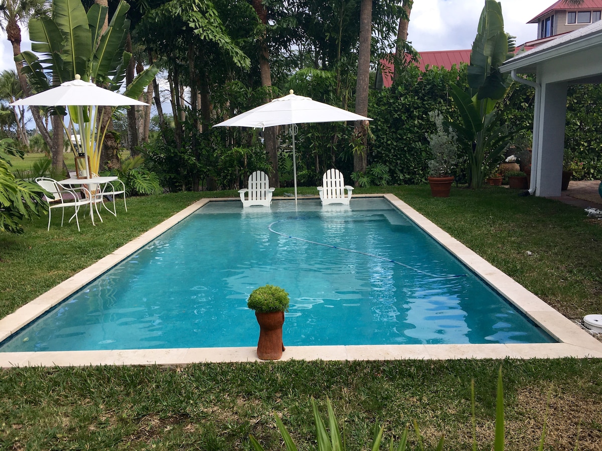 A heated swimming pool is surrounded by lush greenery, featuring two white lounge chairs and two umbrellas providing shade. A small potted plant is positioned at the center of the pool, contributing to the serene outdoor environment.