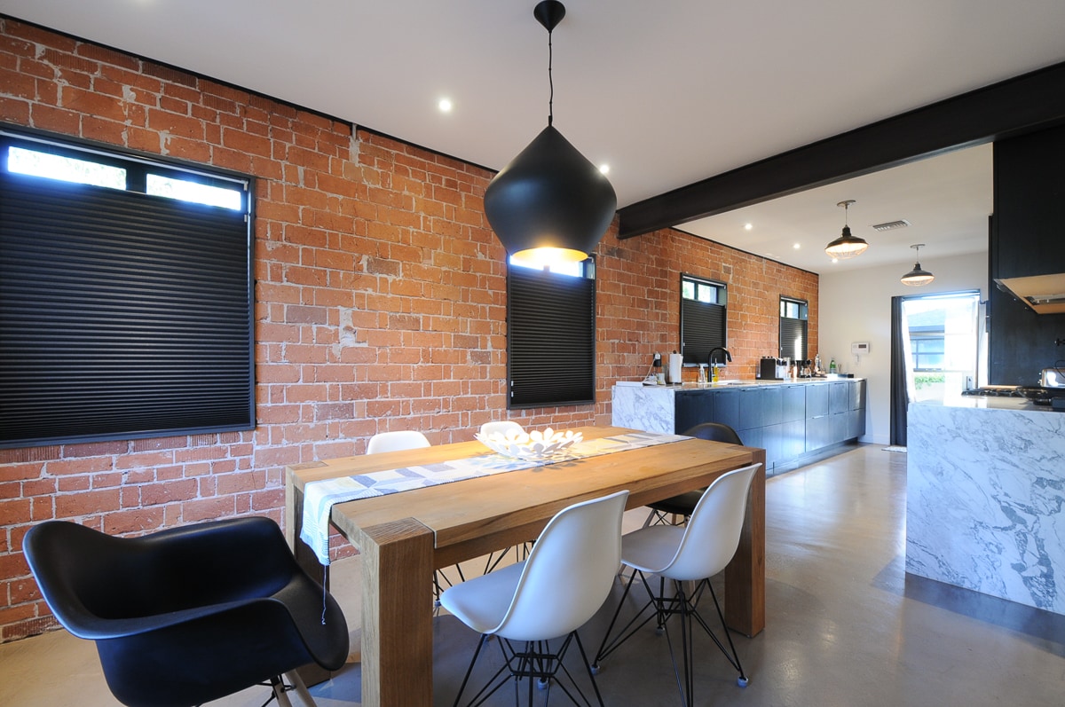 A modern kitchen and dining area feature a long wooden dining table surrounded by white chairs. Exposed brick walls and large windows enhance the industrial design. A sleek kitchen island with marble counters is visible in the background, providing additional cooking space.