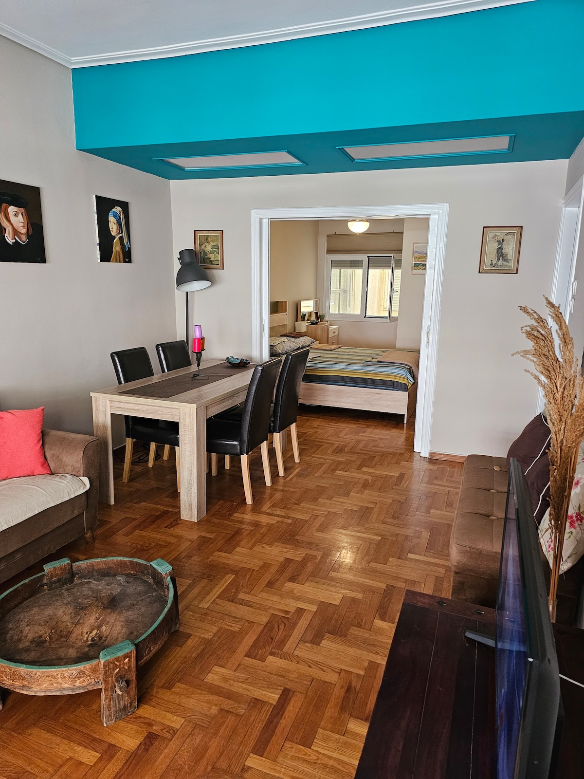 An open living space features a dining table with black chairs set against a light wood floor. A cozy couch adorned with a pink cushion is positioned nearby, and a coffee table rests at the center. An adjacent bedroom with a blue-striped bedspread is visible through the doorway.