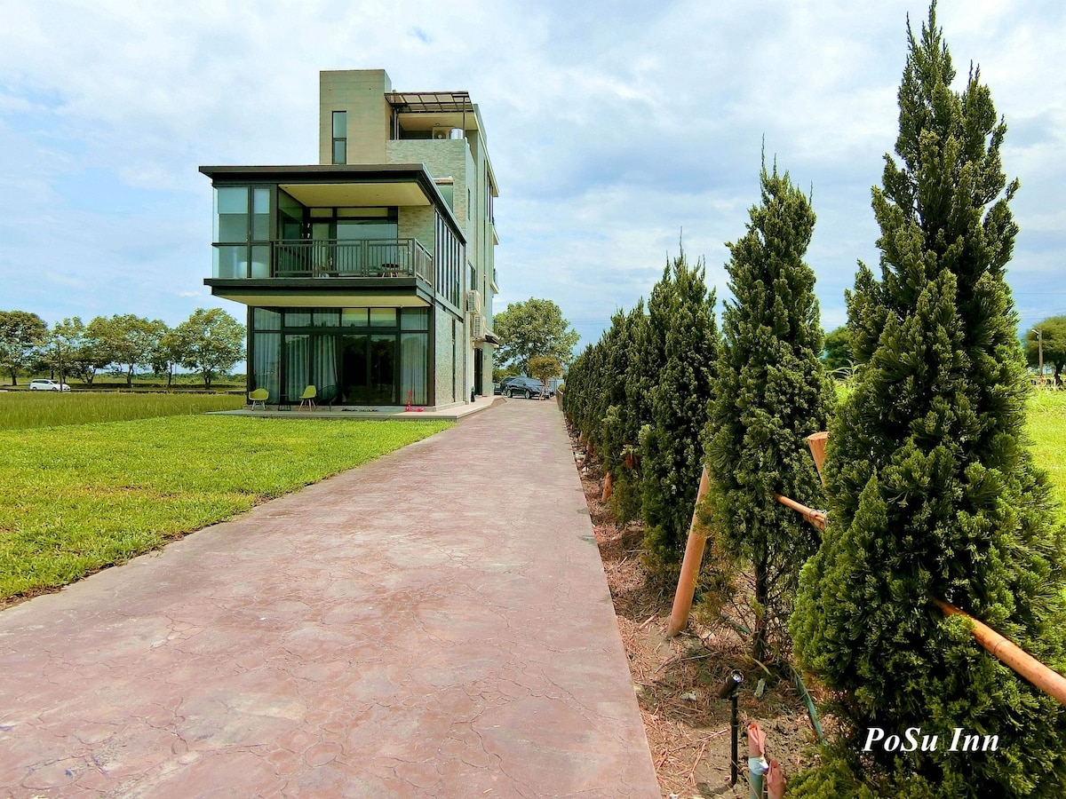A modern building with large glass windows stands next to a neatly paved pathway. Lush green grass surrounds the property, while neatly trimmed trees line the walkway, creating a tranquil and inviting environment.