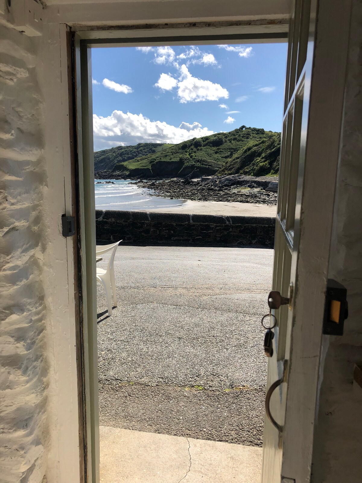 A door stands open, revealing a view of the coastline beyond. The ocean glistens under a clear sky, with gentle waves lapping at the shore. Lush greenery is visible on the opposite hillside, enhancing the tranquil outdoor scene.