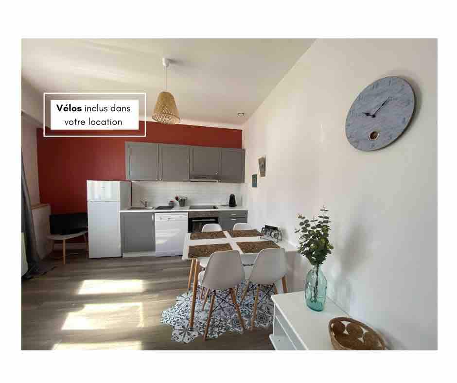 A well-equipped kitchen is shown, featuring modern appliances including a refrigerator and dishwasher. A dining table with four chairs is positioned under a decorative lamp. A clock is mounted on the wall, and a plant adds a touch of greenery to the space.