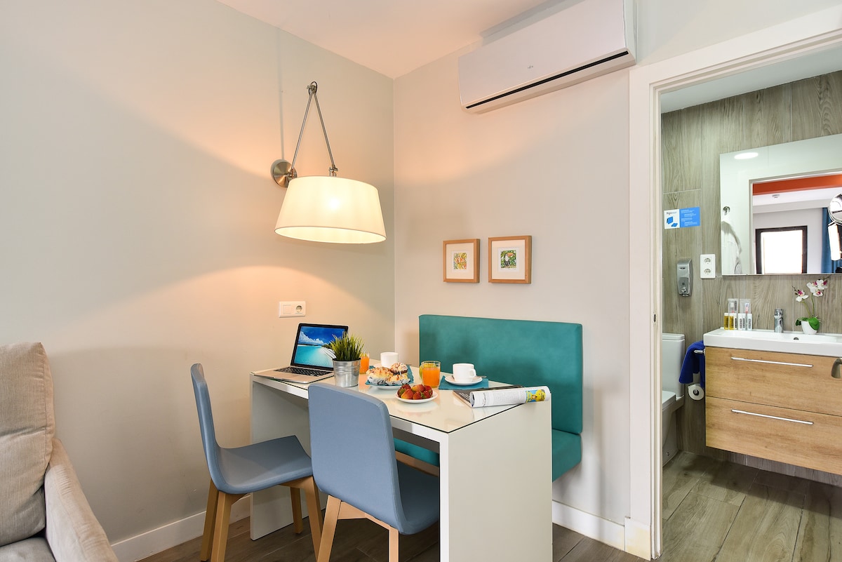 A compact dining area is set with a small table and two chairs, complemented by a laptop on the table. A light fixture hangs above, illuminating the space. In the background, a modern bathroom is partially visible, with wooden accents and amenities.