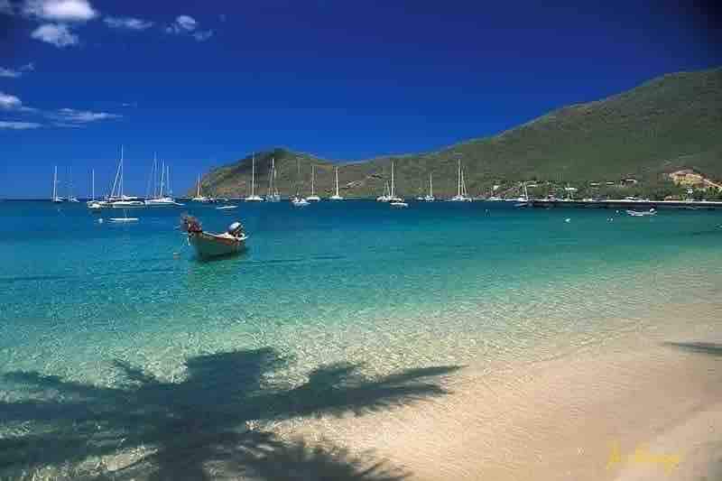 A serene beach scene features calm turquoise waters and a sandy shore. In the foreground, a wooden boat is anchored, with a distant view of sailing vessels. Lush green hills rise in the background under a clear blue sky dotted with a few clouds.