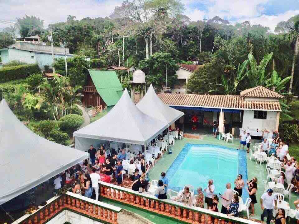 A spacious outdoor area is shown, featuring a clear pool surrounded by seating and tents. Groups of people are gathered, and lush greenery from the surrounding landscape is visible, providing a natural backdrop to the event setting.