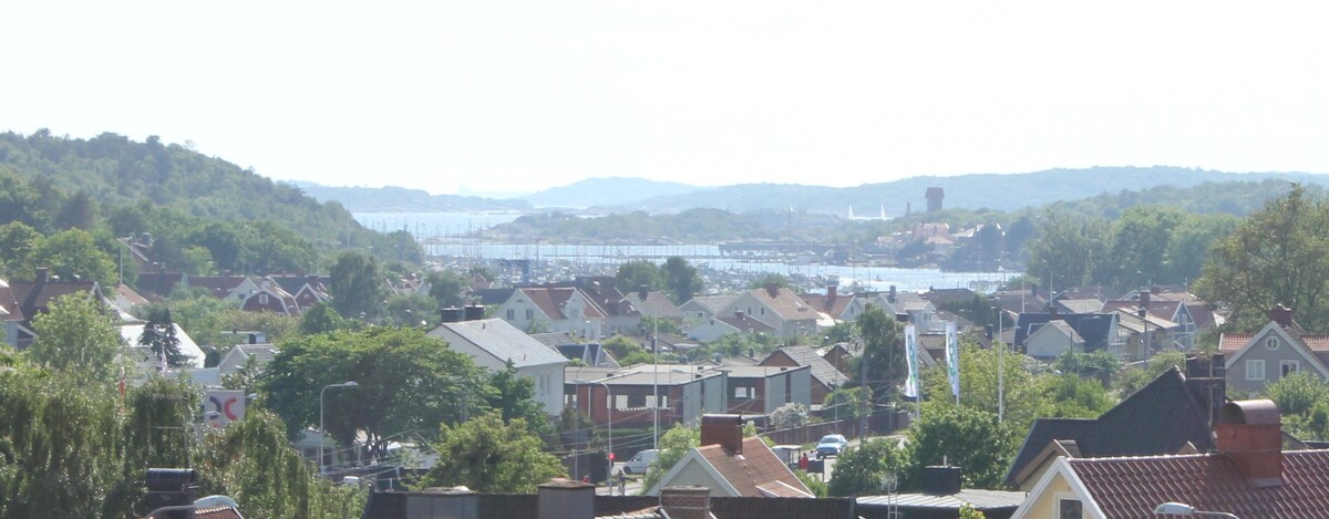 A panoramic view of the surrounding area showcases a mix of residential buildings and greenery, leading towards a distant shoreline. The sea extends into the background, with gentle hills rising along the coastline under a clear sky.