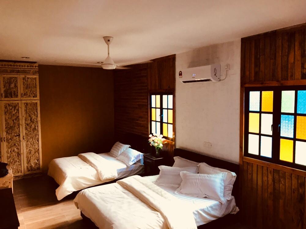 A spacious room is equipped with two comfortable beds, each layered with clean linens. Natural light filters through colorful window panes, enhancing the wooden wall accents. A wardrobe is positioned against one wall, accompanied by a ceiling fan for comfort.