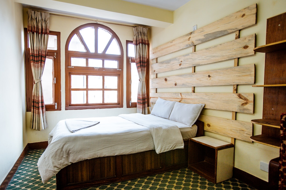 A well-lit guest room is featured, with a spacious bed positioned against a wooden accent wall. Natural light pours in through a set of large arched windows, framed by light curtains. A bedside table and bookshelves are included in the room.