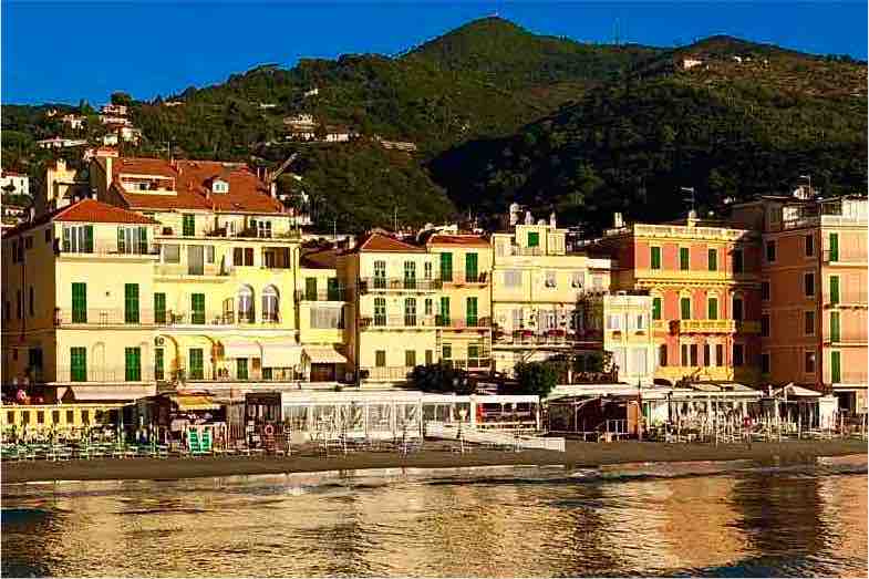 A coastal view of a charming waterfront, highlighting colorful buildings with green shutters lining the shore. The image captures the vibrant atmosphere of the area, complemented by a softly rippling sea and a backdrop of lush hills under clear blue skies.