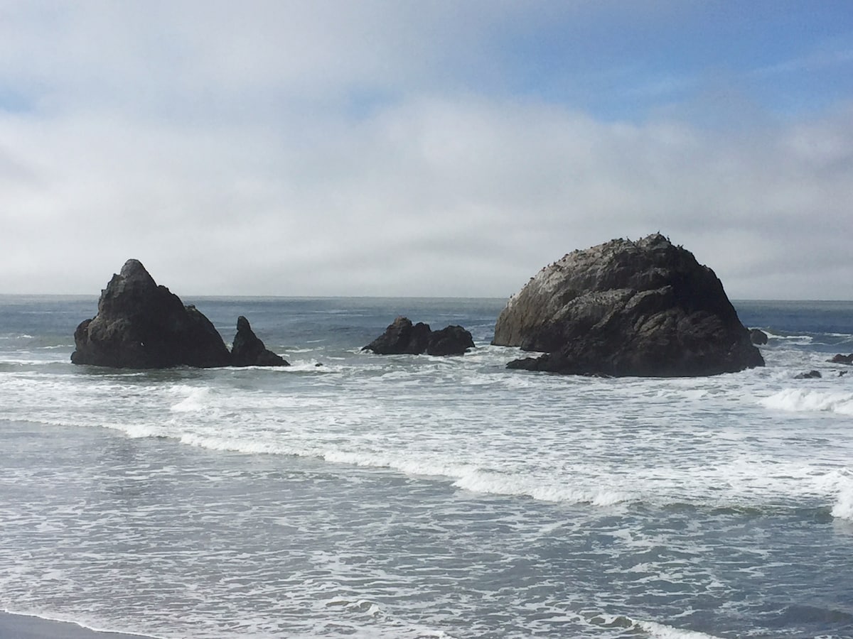 Two rocky formations rise from the ocean, surrounded by gentle waves lapping at the shore. The water reflects a light blue hue, and a soft blanket of clouds drifts across a bright sky, creating a serene coastal scene.